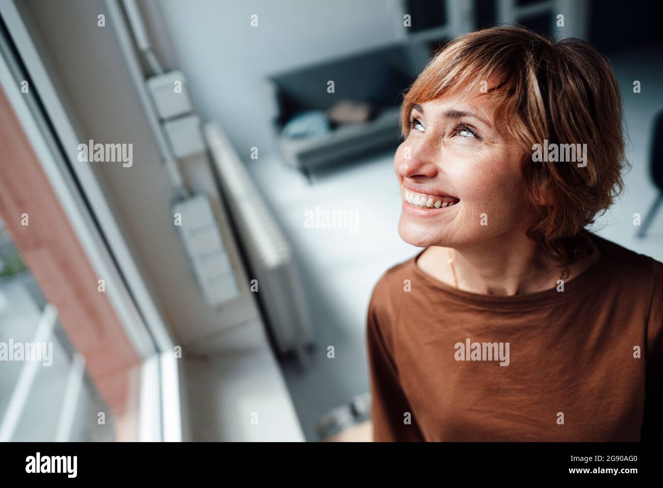 Geschäftsfrau lächelt, während sie am Fenster im Büro aufschaut Stockfoto