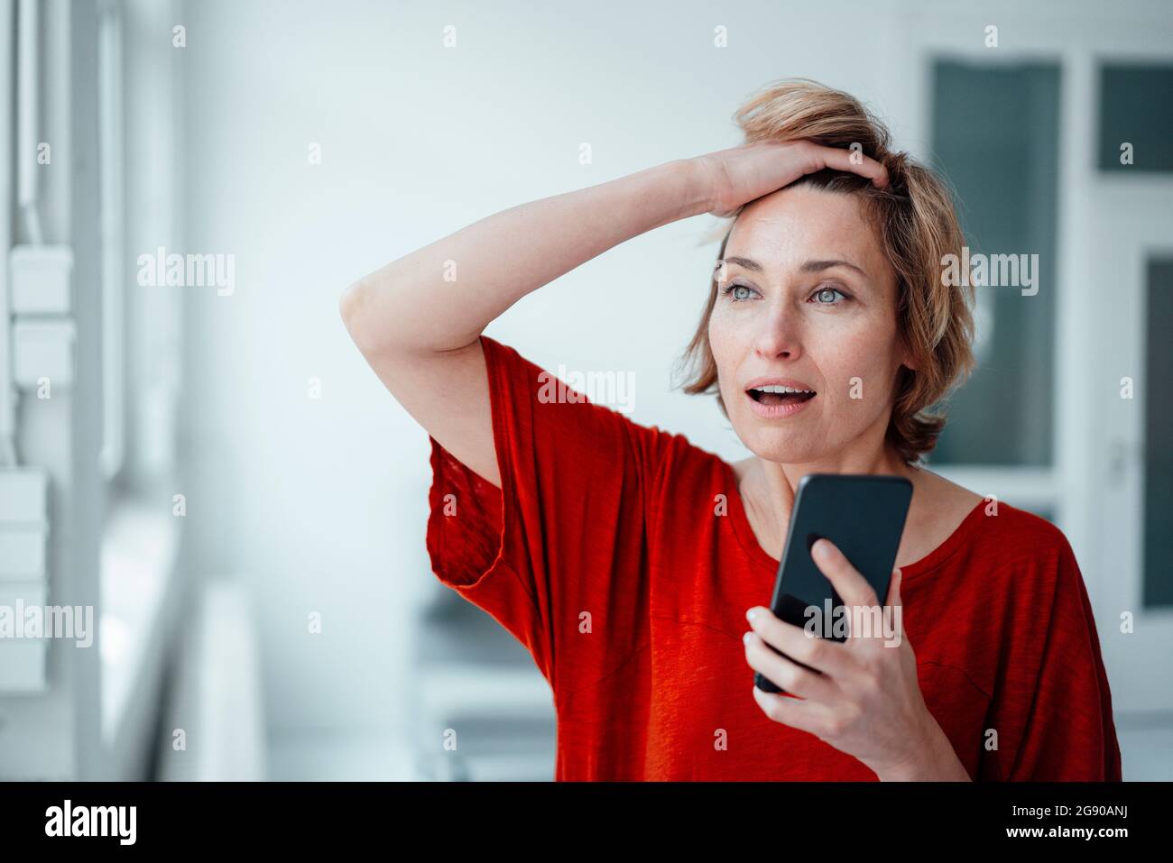 Geschäftsfrau mit offenem Mund und Hand im Haar mit Smartphone im Büro Stockfoto