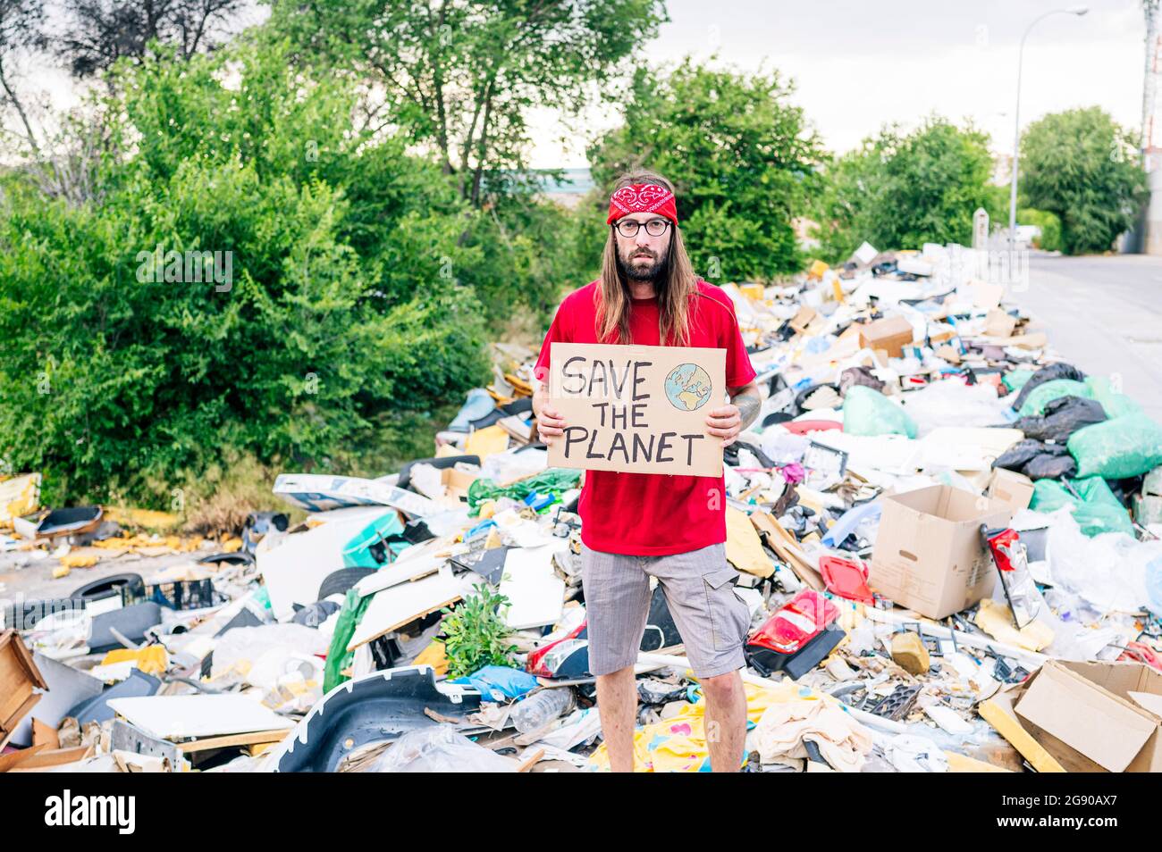 Junger Hippie-Mann hält den Planeten Karton, während er auf Müllhalde steht Stockfoto