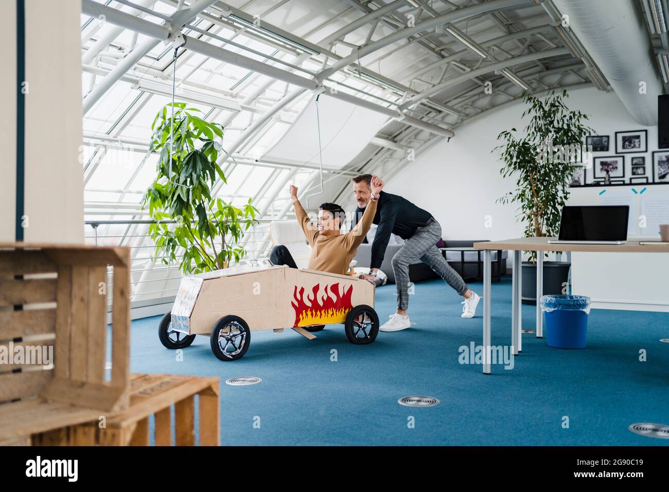 Kollege schiebt fröhlichen Geschäftsmann im Spielzeugauto im kreativen Büro sitzen Stockfoto