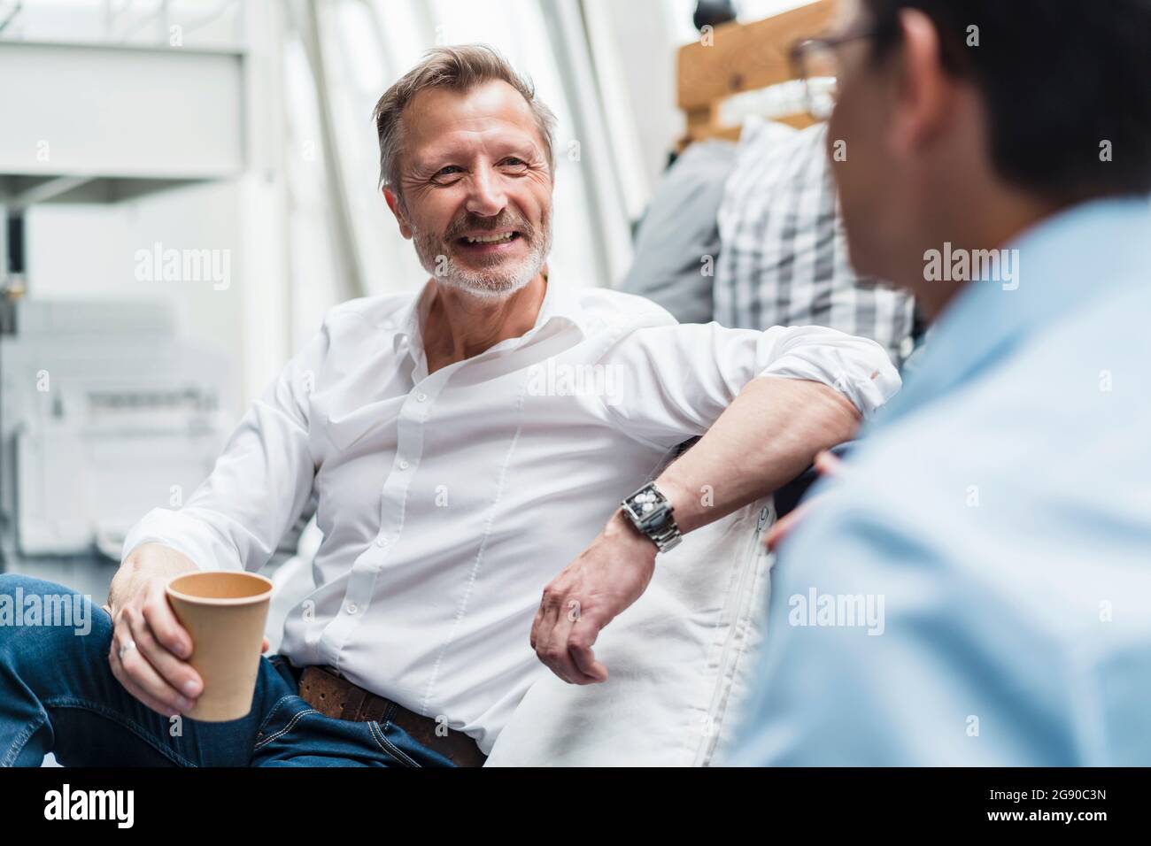 Lächelnder, männlicher Profi, der einen Einweg-Becher hält, während er mit einem Kollegen im Büro sitzt Stockfoto Lächelnder, männlicher Profi, der einen Einweg-Becher hält, während er mit einem Kollegen im Büro sitzt Stockfoto