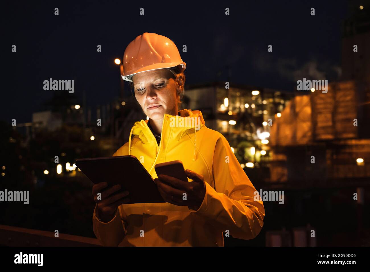 Weibliche Profi in gelbem Hardhut und Regenmantel mit drahtlosen Technologien in der Nacht Stockfoto