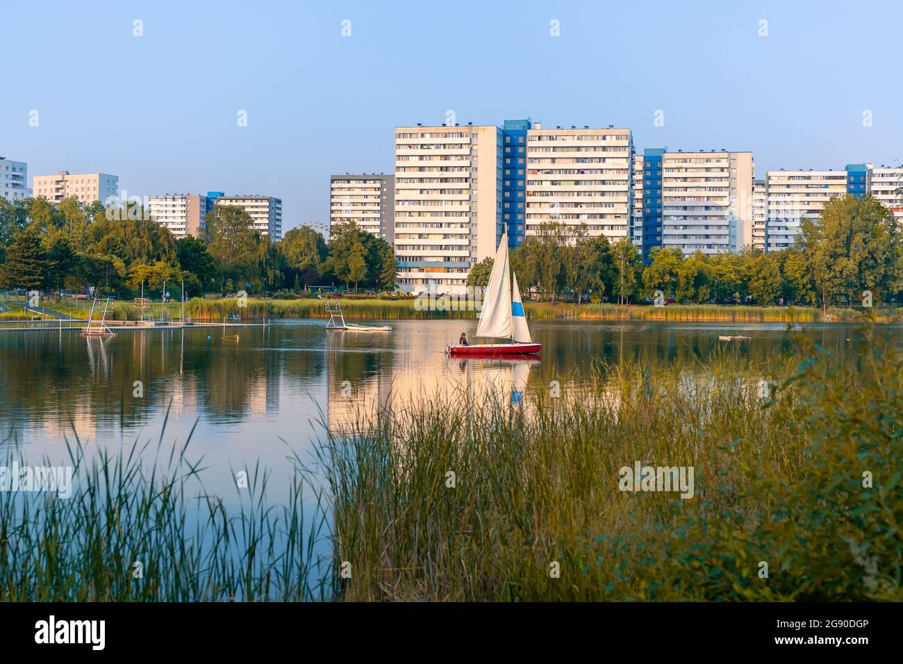 Rotes Segelboot segelt in dem kleinen See, umgeben von Bäumen und Sträuchern. Hohe Wohngebäude im Hintergrund. Katowice, Schlesien, Polen. Stockfoto