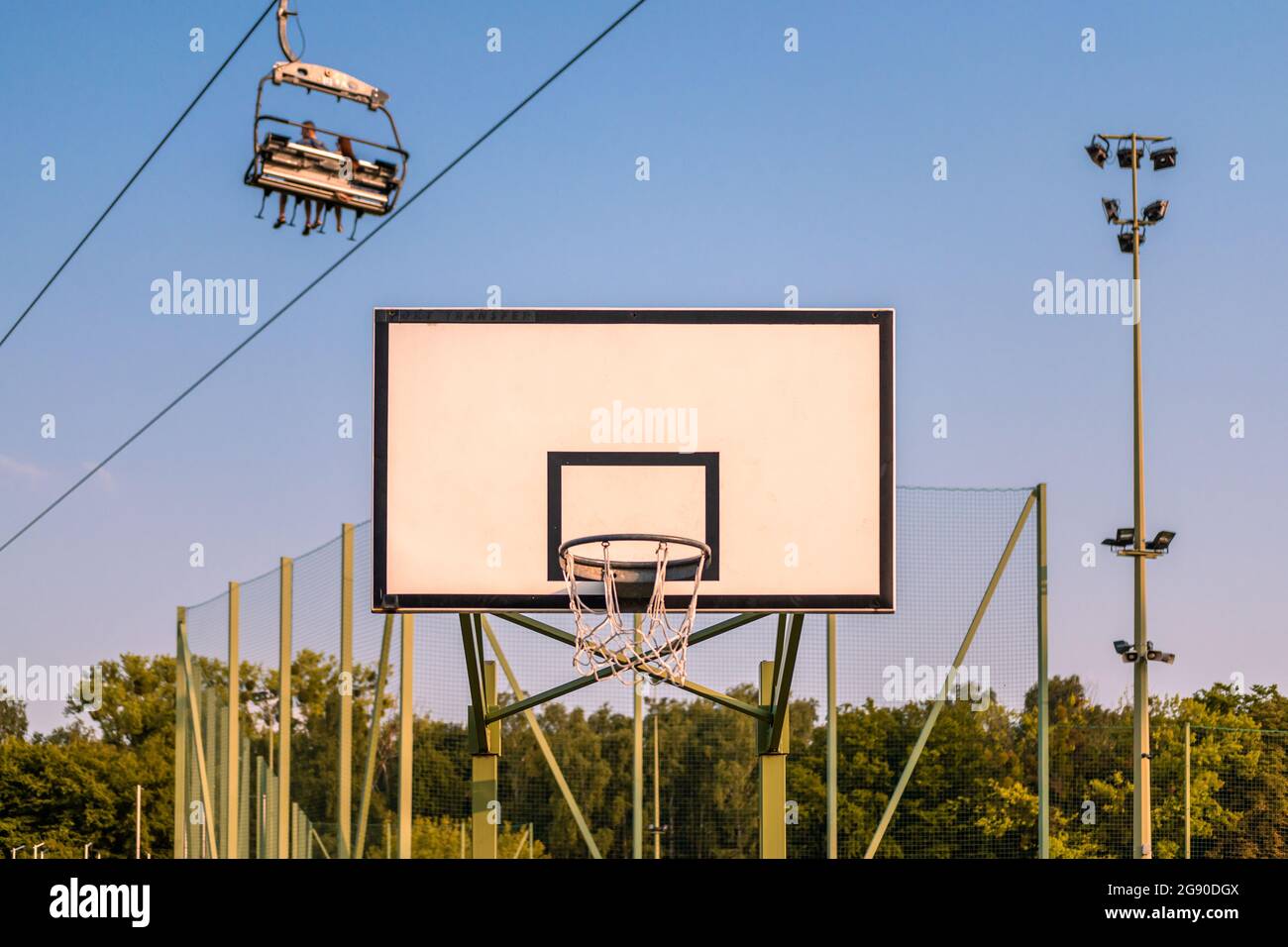 Basketballbrett gegen Baumspitzen, blauer Himmel und viersitziger Sessellift mit drei Personen darin. Stockfoto