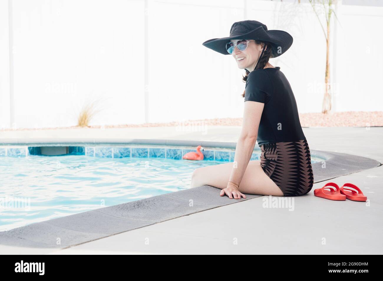 Lächelnde Frau mit Sonnenbrille, die im Urlaub am Pool sitzt und wegschaut Stockfoto