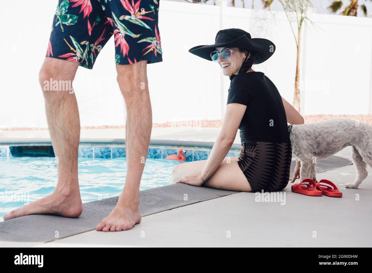 Glückliche Frau mit Hund, die während des Urlaubs hinter einem Mann am Pool sitzt Stockfoto