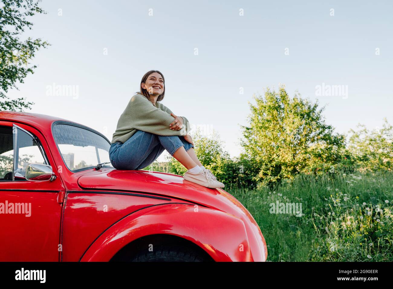 Lächelnde junge Frau, die die Knie umarmt und auf der Motorhaube des Autos sitzt Stockfoto
