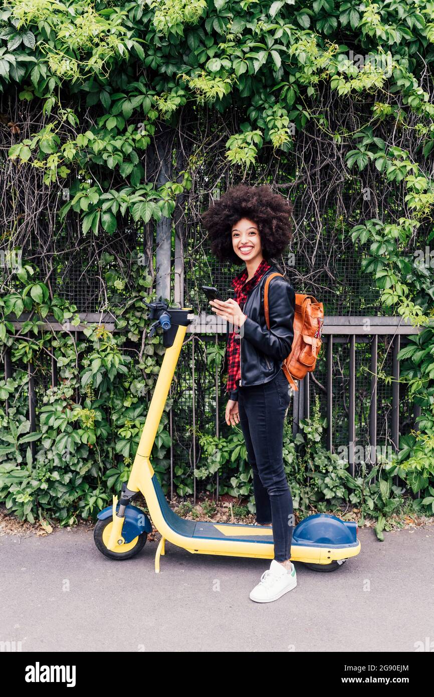 Lächelnde Frau hält Mobiltelefon, während sie mit einem elektrischen Motorroller auf der Straße steht Stockfoto