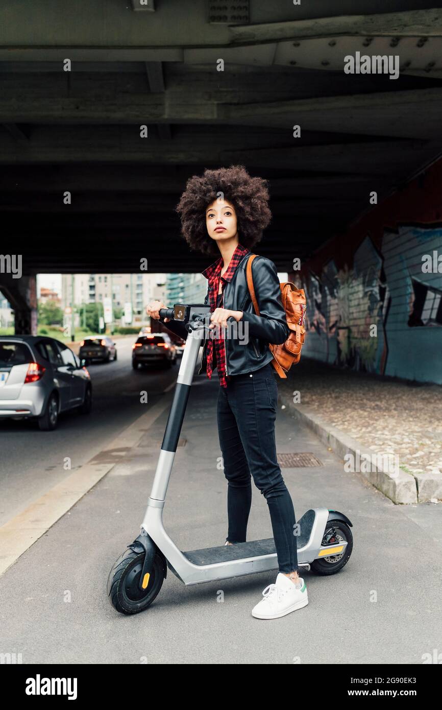 Junge Frau, die mit einem elektrischen Motorroller auf der Straße unter der Brücke steht Stockfoto