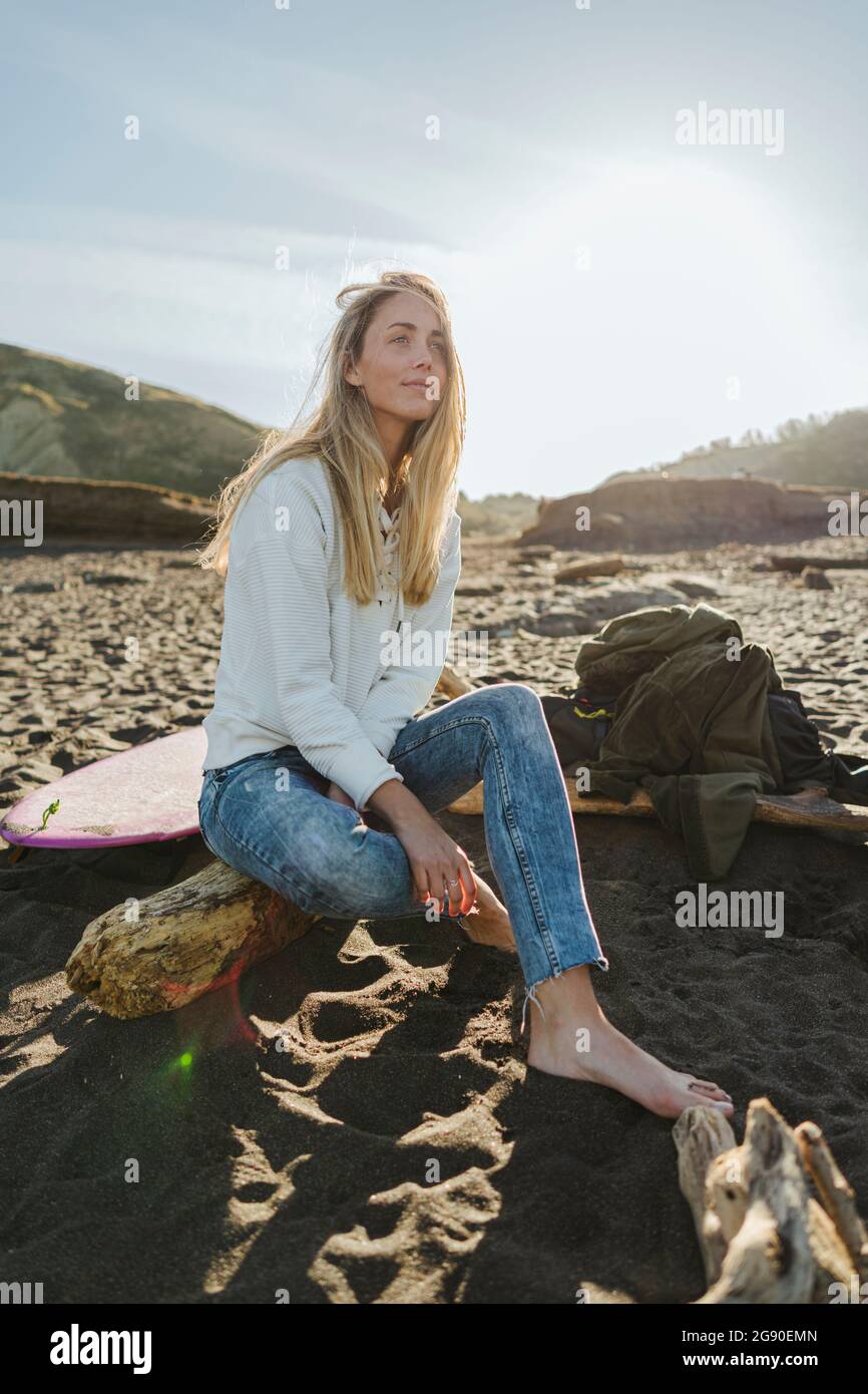 Schöne Frau mit blonden Haaren, die am Strand auf dem Baumstamm sitzt Stockfoto
