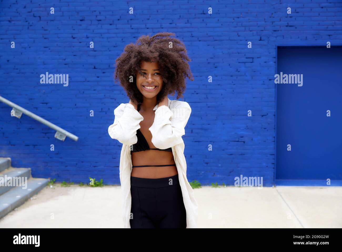 Lächelnde, schöne Afro-Frau, die vor der blauen Wand wegschaut Stockfoto