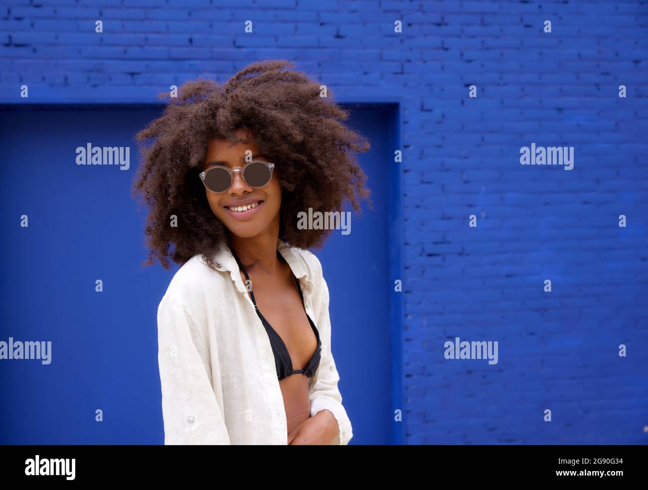 Lächelnde junge Afro-Frau mit Sonnenbrille vor der blauen Wand Stockfoto