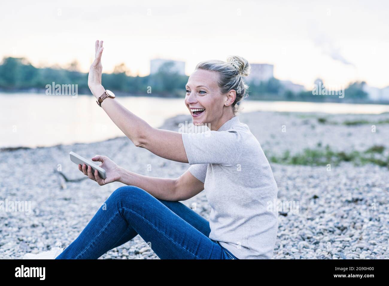 Fröhliche Frau winkt, während sie mit dem Mobiltelefon am Flussufer sitzt Stockfoto