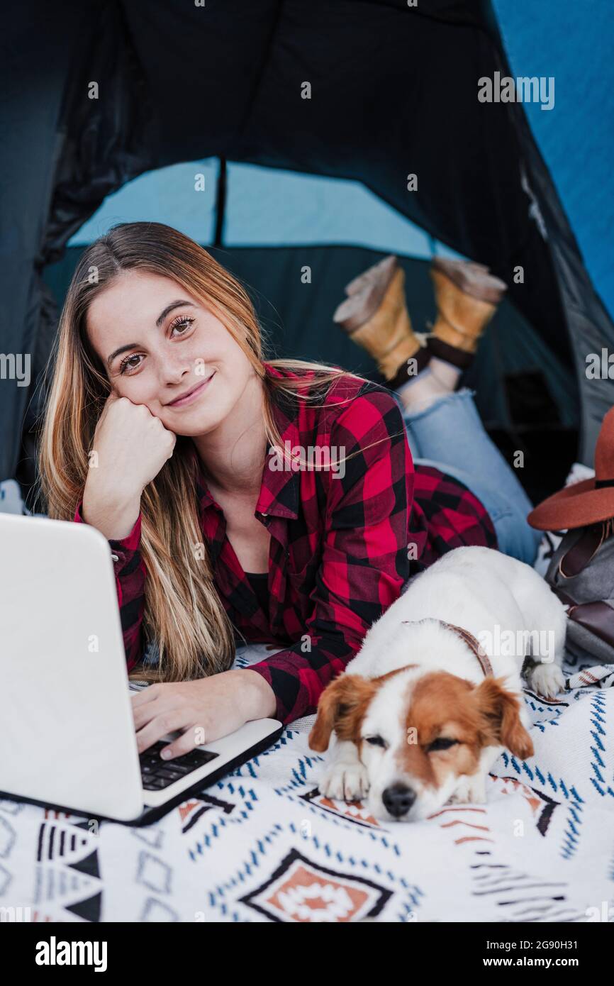 Lächelnd schöner junger digitaler Nomade, der mit Laptop und Hund im Zelt liegt Stockfoto