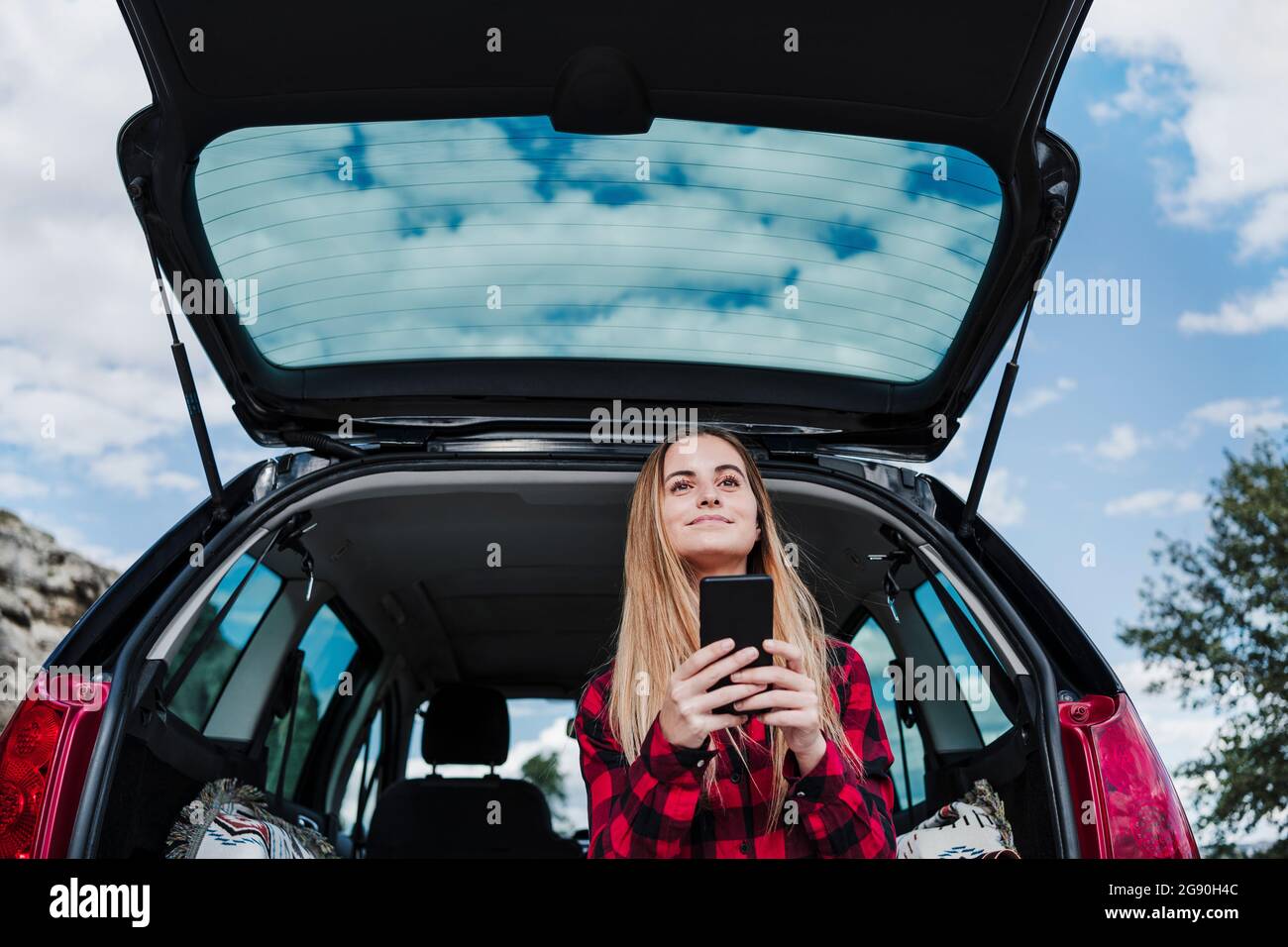 Blonde Frau, die mit einem Smartphone im Kofferraum sitzt Stockfoto