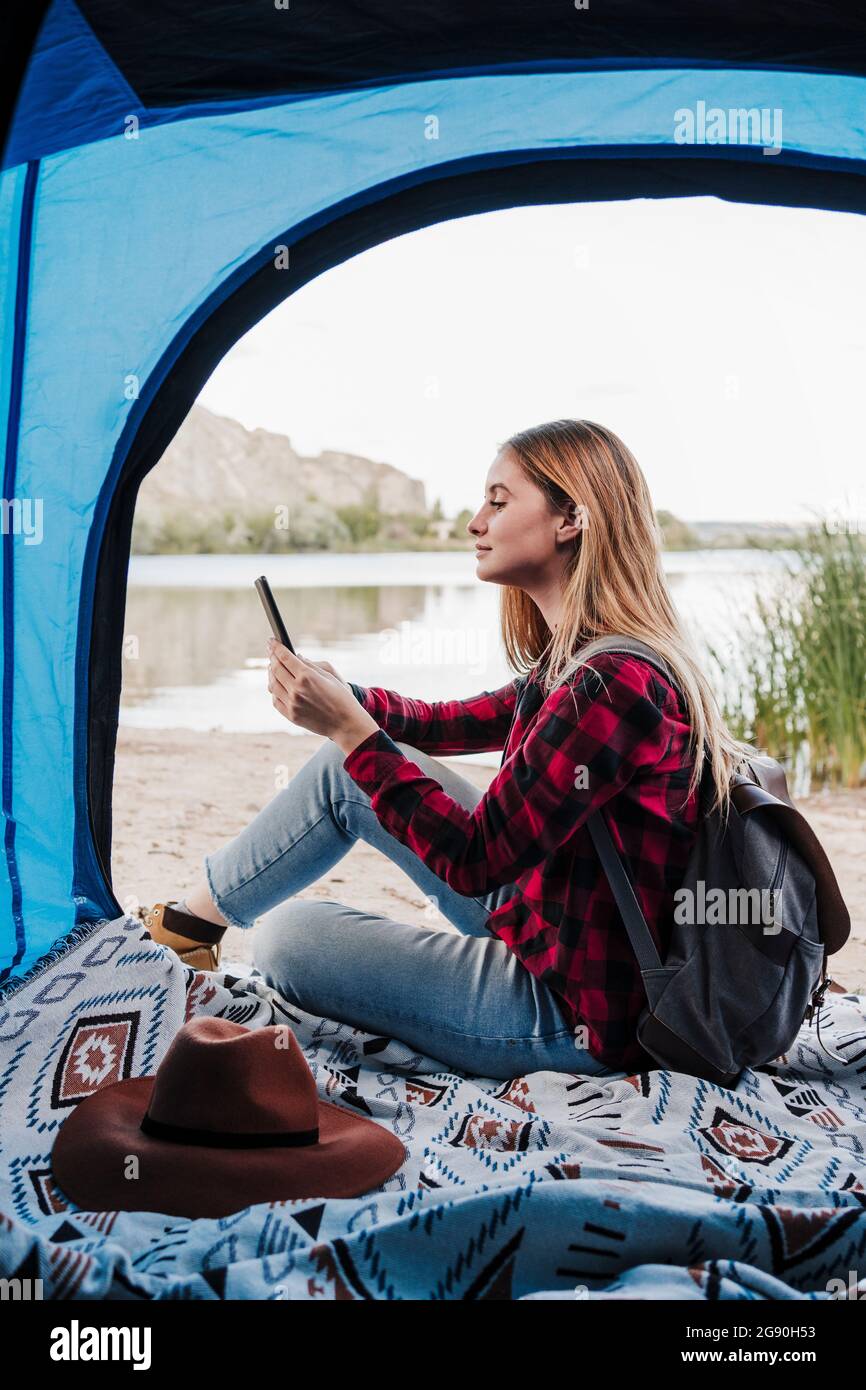 Schöne junge blonde Frau, die im Zelt über ein Smartphone im Internet surft Stockfoto