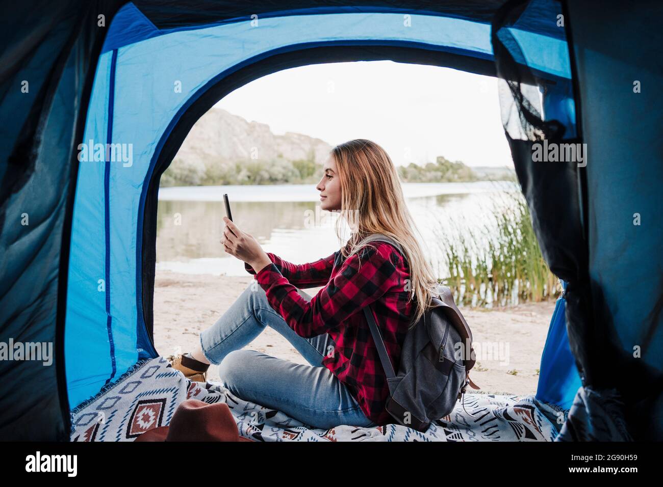 Junge blonde Frau SMS-Nachrichten über Smartphone, während sie im Zelt sitzt Stockfoto
