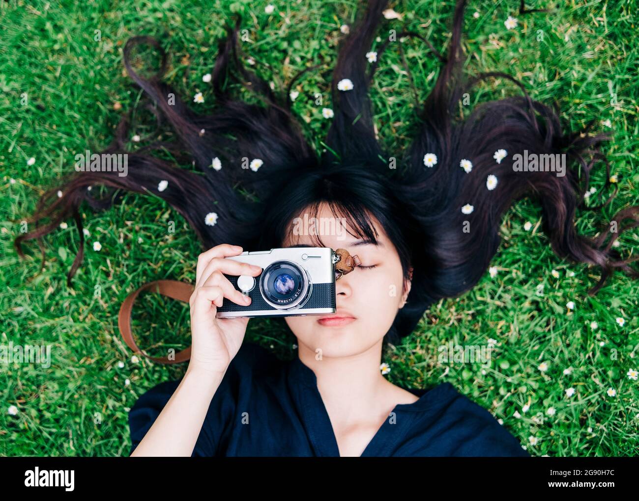 Fotografin mit Blumen im Haar, die die Kamera halten, während sie auf dem Gras liegt Stockfoto