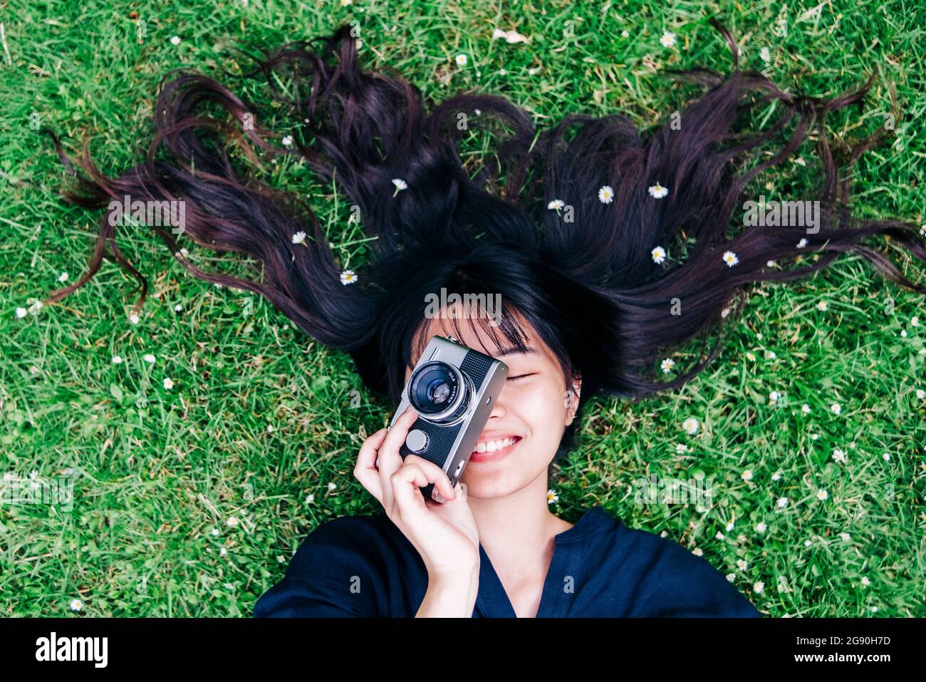 Glückliche Fotografin mit Blumen im Haar, die die Kamera halten, während sie auf dem Gras liegt Stockfoto