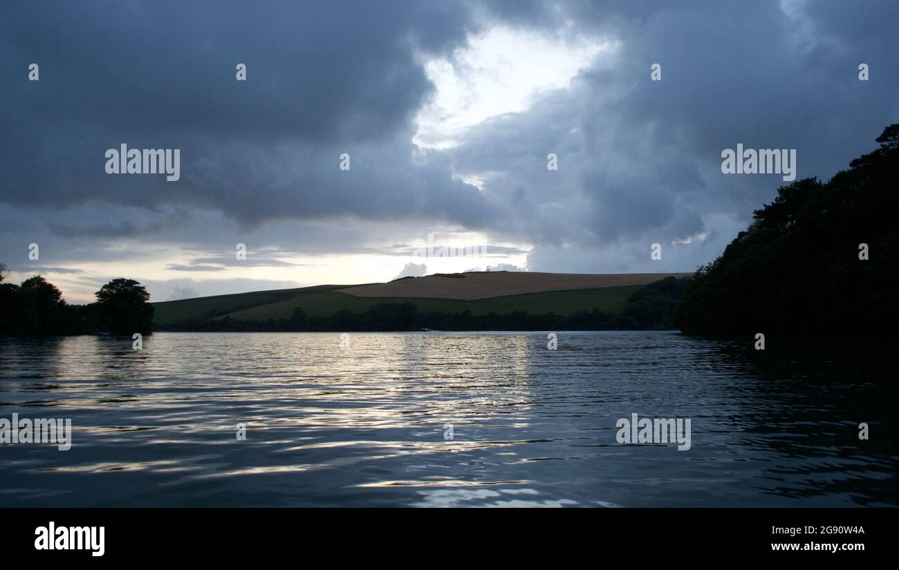 Blick auf die Küste in Twilight Blue: Ruhiges Wasser, Ackerland auf dem Hügel im Hintergrund, dunkle schattige Bäume auf beiden Seiten des Sees oder der Mündung unter dem Stockfoto