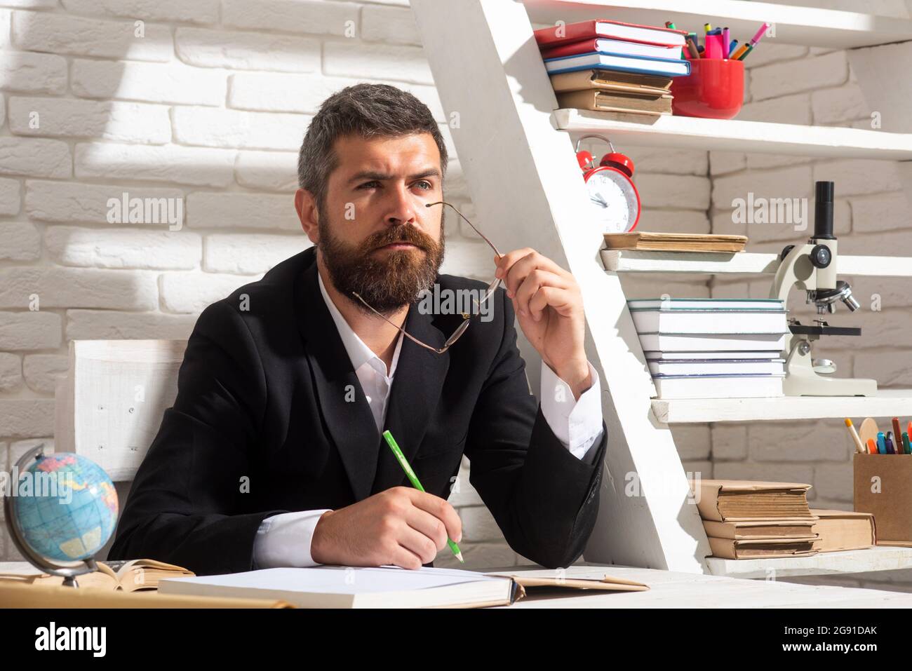 Porträt eines denkenden männlichen Lehrers im Klassenzimmer. Der Lehrer sitzt auf dem Schreibtisch und schreibt Text in einem Buch im Klassenzimmer Stockfoto