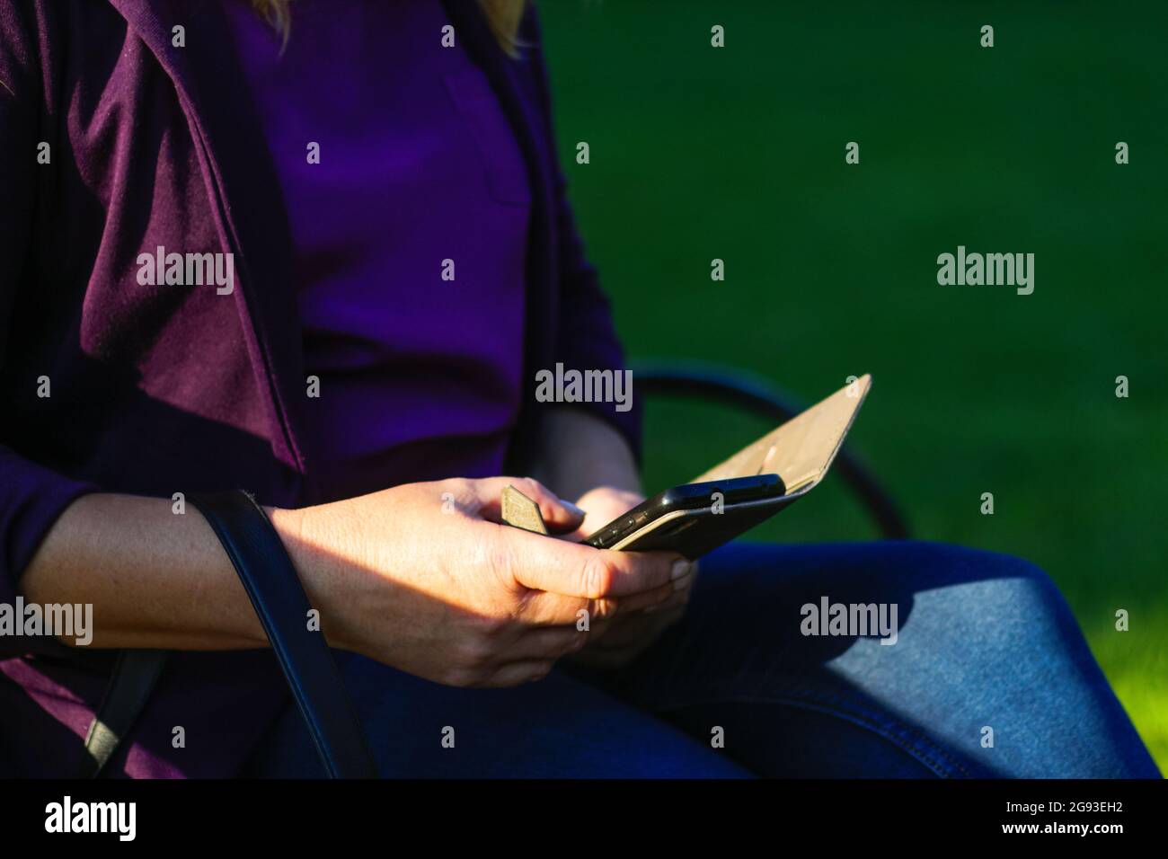 Unschärfe kaukasische blonde Frau sucht auf dem Telefon draußen, im Freien. Erwachsene Frau in lila Bluse im Park mit Smartphone, während sie auf einer Bank sitzt Stockfoto