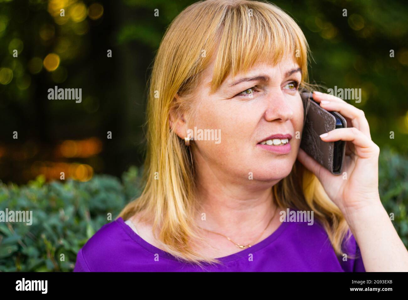 Unschärfe-Nahaufnahme lächelnde kaukasische blonde Frau, die im Freien telefoniert. 40-jährige Frau in lila Bluse im Park. Erwachsener Stockfoto