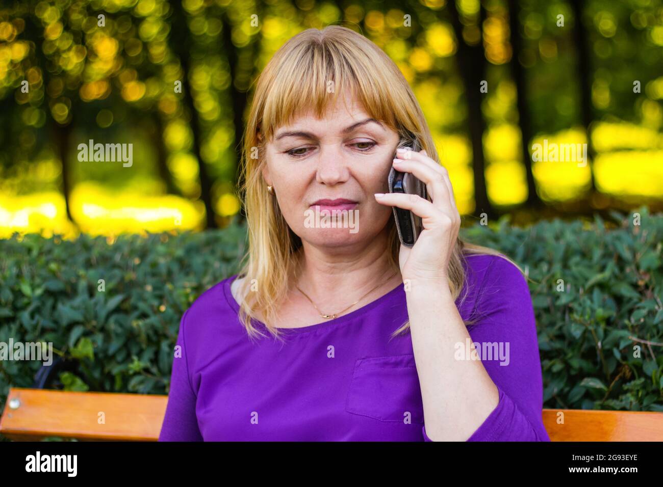 Unschärfe-Eleganz kaukasische blonde Frau reden, sprechen auf dem Telefon draußen, im Freien. 45er Jahre alte Frau in lila Bluse im Park. Erwachsene Frauen Stockfoto