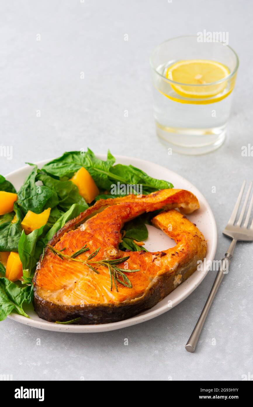 Gebratenes Lachssteak mit Spinat und gelben Tomaten auf einem Teller auf grauem Grund, ein Glas Wasser mit Zitrone, ein herzhaftes Mittagessen mit gesundem rotem Fisch Stockfoto