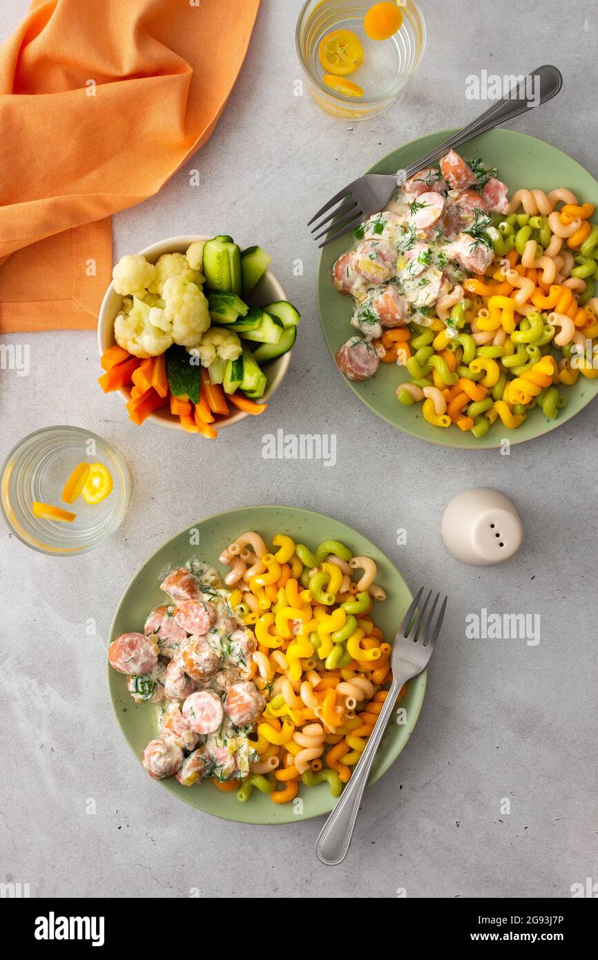 Bunte Pasta mit gekochten Würstchen in saurer Rahmsoße, frischem Gemüse und gekochtem Blumenkohl, Wasser mit Kumquats Stockfoto