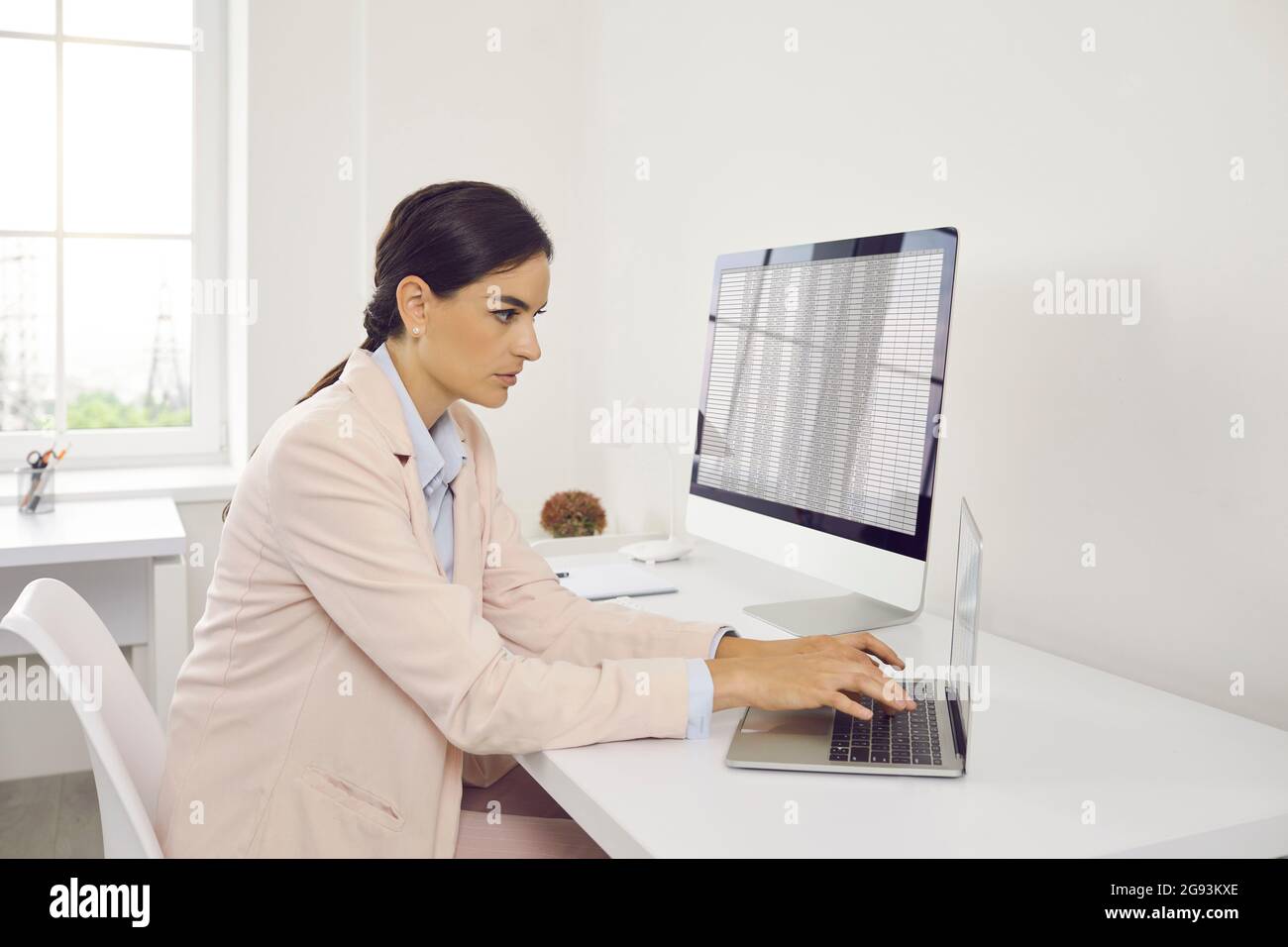 Buchhalterin, die mit einem Laptop arbeitet, während sie an ihrem Schreibtisch mit einem Desktop-Computer sitzt Stockfoto