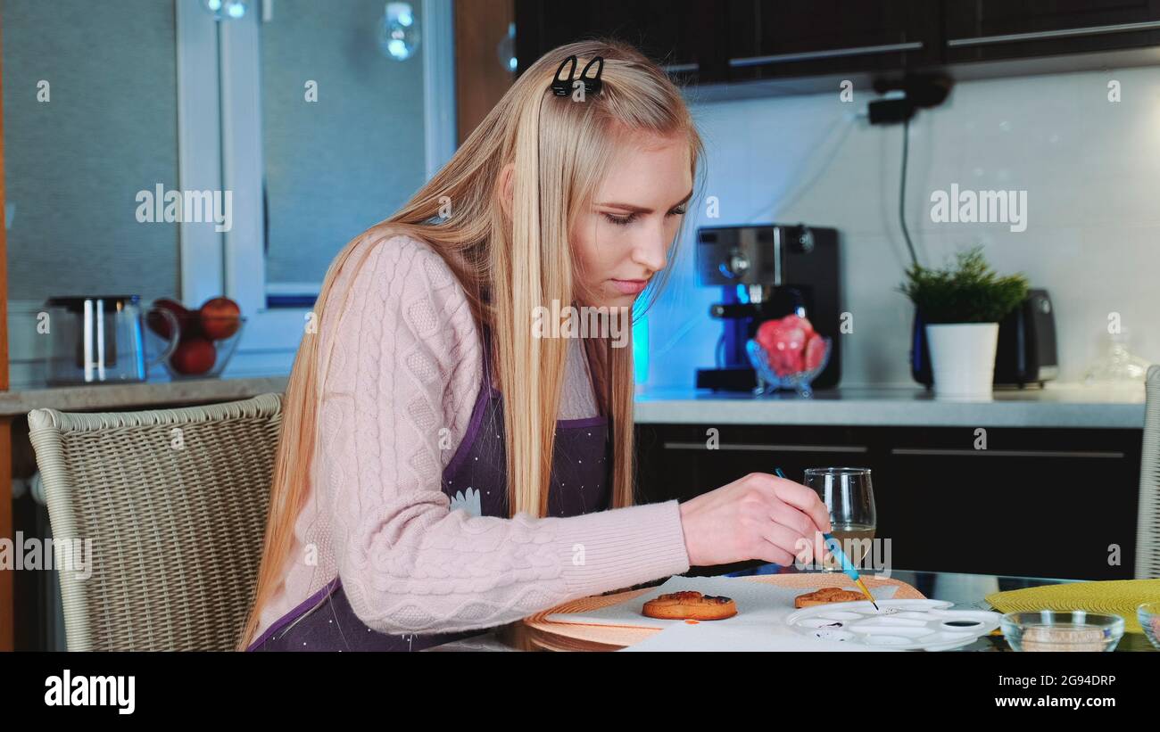 Hübsche junge Frau lernen, Lebkuchenkekse zu Hause zu dekorieren. Sie sitzt am Tisch und malt. Stockfoto