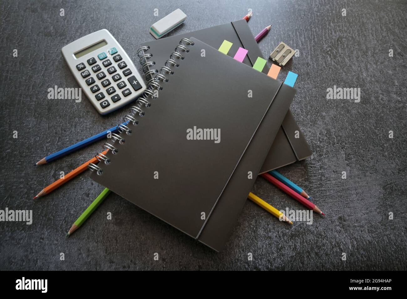 Schwarze Spiralhefte, bunte Buntstifte, Taschenrechner und stationäres Zubehör auf einem dunkelgrauen Schreibtisch, Kopierraum, ausgewählter Fokus, enge Schärfentiefe Stockfoto