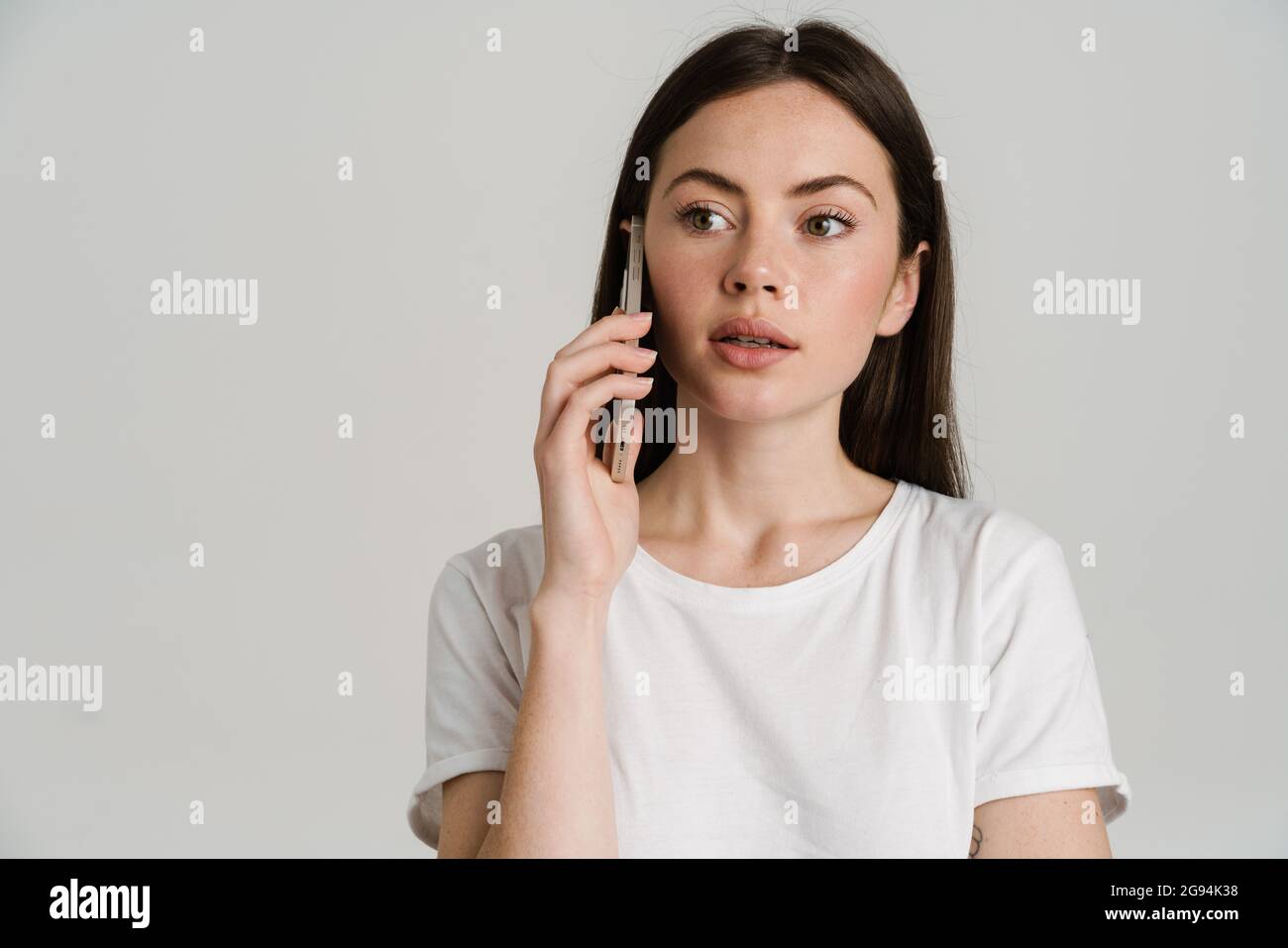 Nahaufnahme Porträt einer jungen weißen Frau mit langen braunen Haaren tragen weißes T-Shirt stehen auf grauem Hintergrund reden auf Handy, das aussagt Stockfoto