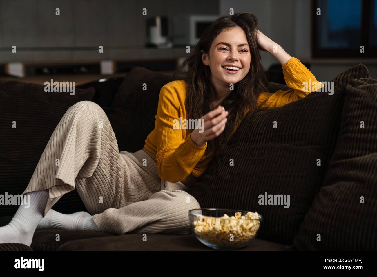Lächelnde junge Brünette Frau, die Popcorn isst, während sie zu Hause auf einer Couch sitzt Stockfoto