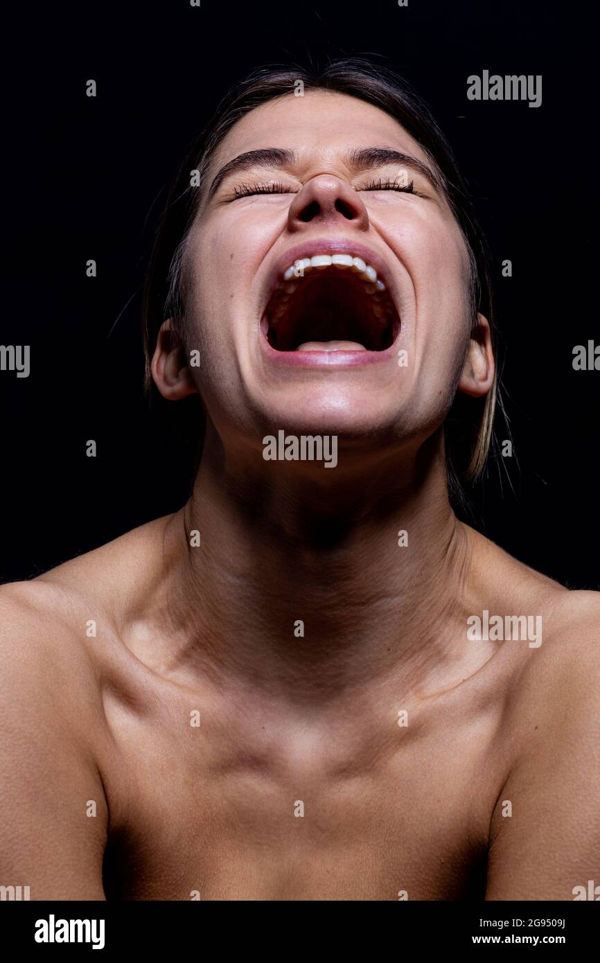 Schreiende Frau auf schwarzem Hintergrund. Das Gesicht einer jungen Frau mit offenem Mund, während sie schreit, isoliert von Schwarz. Sie ist frustriert und leidet unter emotionalen Schmerzen. Sie ist stressig und verrückt. Stockfoto
