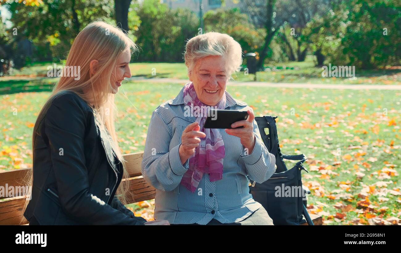 Ältere Großmutter, die ihre Enkelin auf dem Smartphone zeigt. Sie sitzen auf der Bank und reden im Herbstpark. Stockfoto
