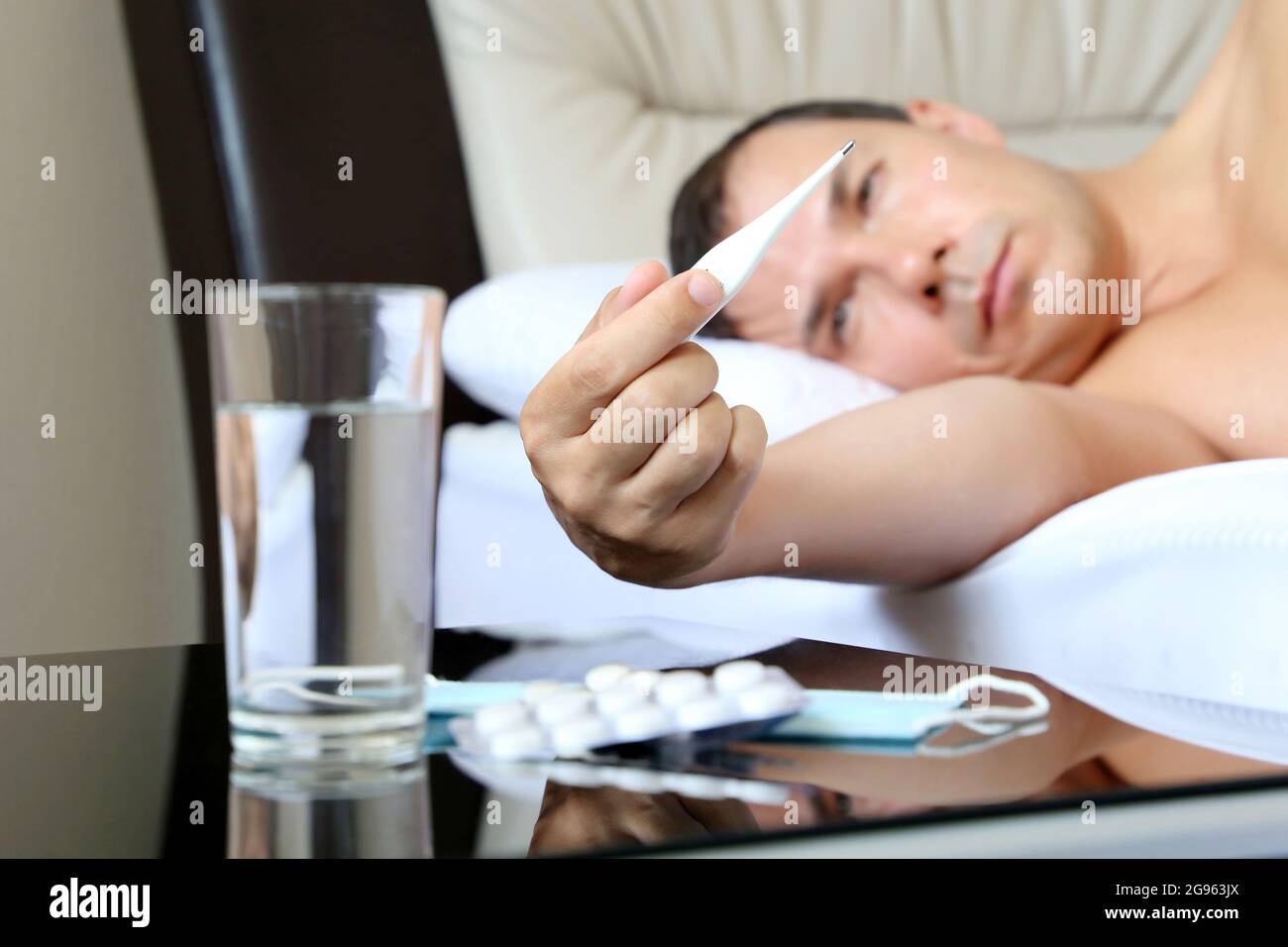 Kranker Mann liegt mit Digitalthermometer im Bett, Pillen und Wasserglas im Vordergrund. Konzept von Krankheit, Fieber, Coronavirus-Symptome Stockfoto