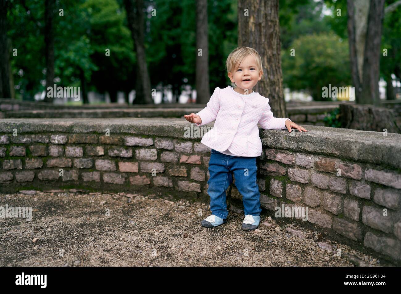 Das kleine Mädchen steht in der Nähe eines niedrigen Steinzauns in einem grünen Park Stockfoto