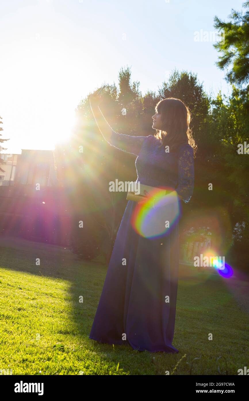 Nettes Mädchen in elegantem Kleid unter Selfie im Sommerpark mit Sonneneinstrahlung, selektiver Fokus Stockfoto