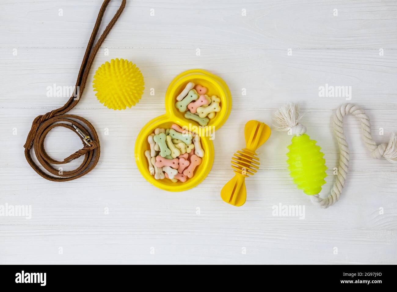 Gelbe Schale mit Snacks, Ball, Leine, ein Knochenspielzeug für Hunde auf weißem Holztisch. Set von Zubehör für Haustiere auf Holz. Nahaufnahme Stockfoto