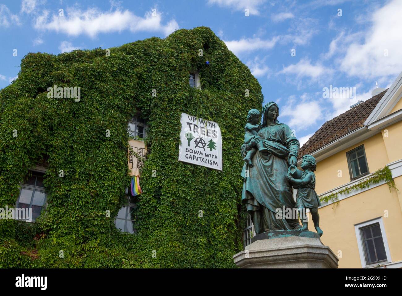 Mutter mit Kindern Statue (Donndorfbrunnen-Statue) und Haus mit 'hoch mit Bäumen runter mit Kapitalismus' Schild in Weimar, Deutschland Stockfoto
