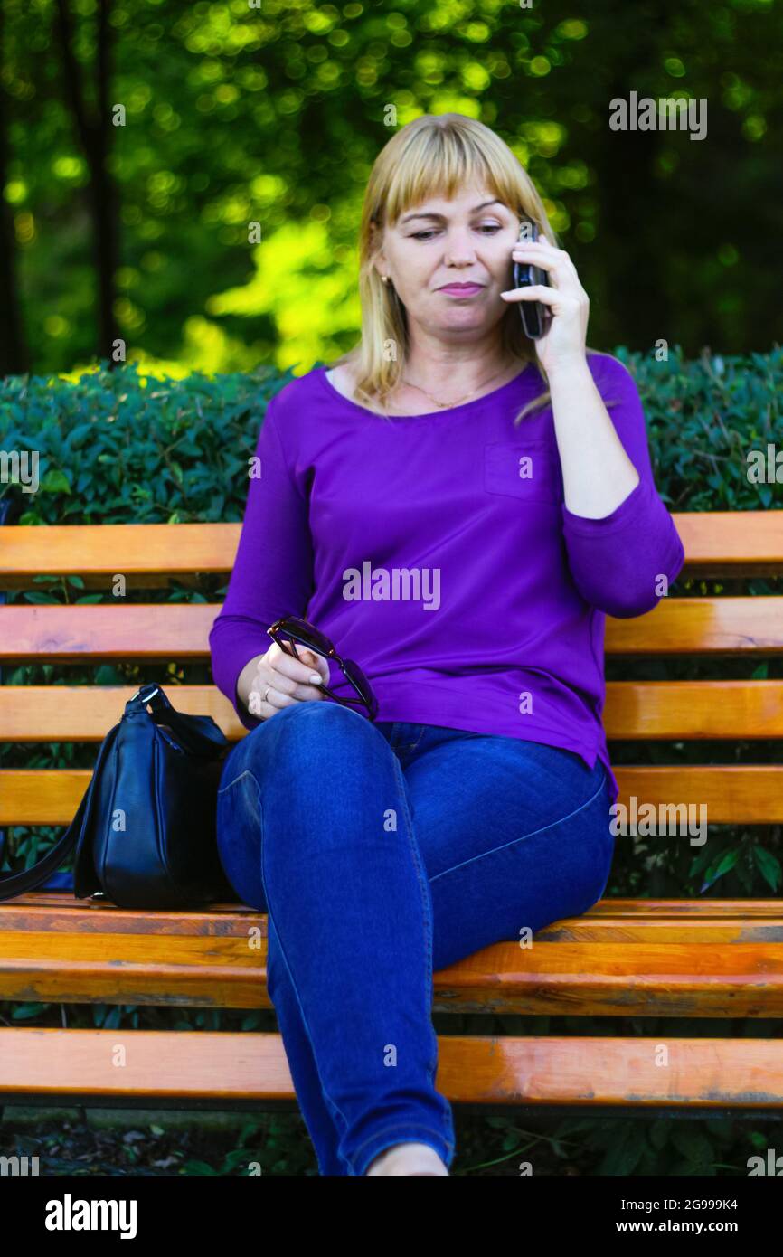 Unschärfe kaukasisch blonde Frau reden, sprechen am Telefon draußen, im Freien. 40-jährige Frau in lila Bluse im Park. Erwachsene Person, die das Telefon benutzt Stockfoto