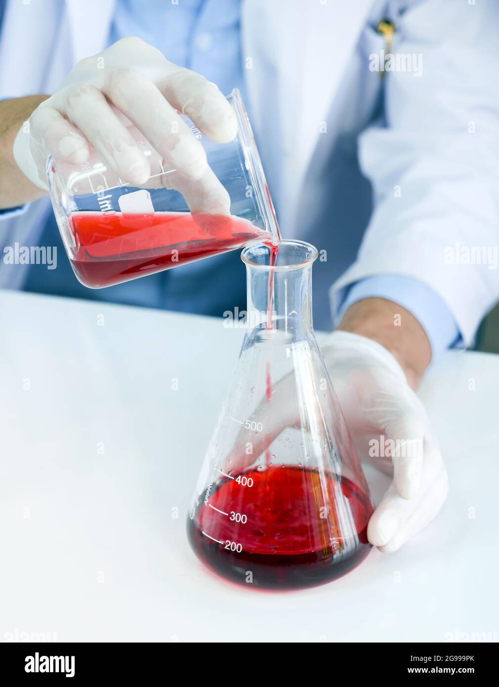 Wissenschaftler im Gewand und Schutzhandschuh mischen vorsichtig rote giftige Chemikalien aus Becher und Erlenmeyerkolben. Stockfoto