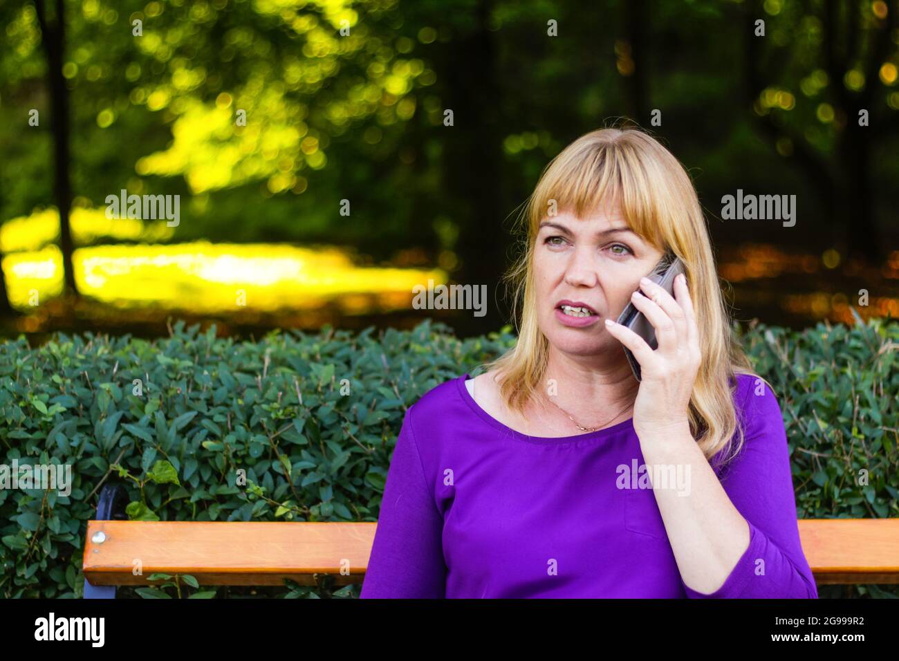 Unschärfe ernsthafte kaukasische blonde Frau sprechen, sprechen am Telefon draußen, im Freien. 40-jährige Frau in lila Bluse im Park auf der Bank. Erwachsener Stockfoto
