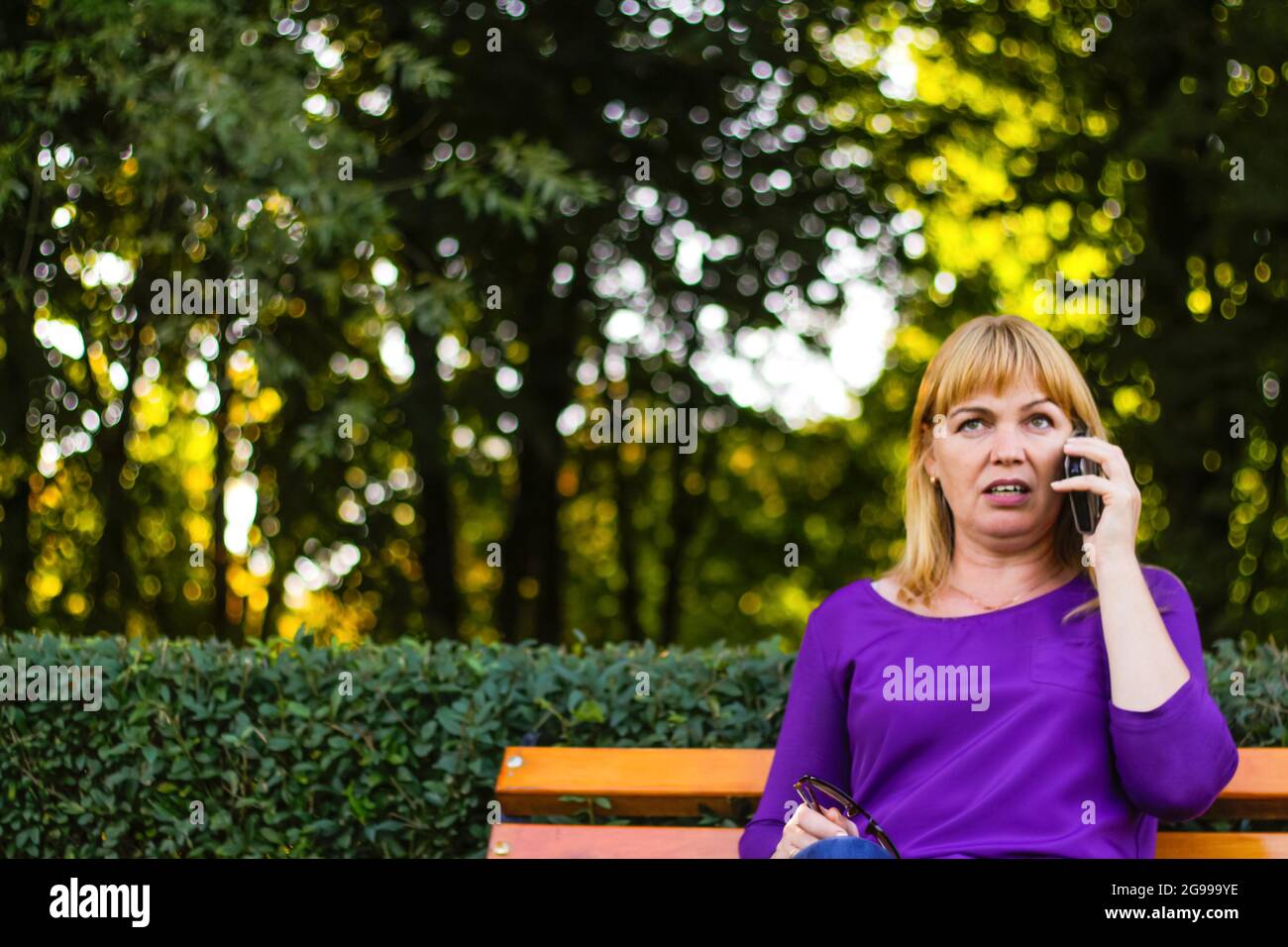 Unschärfe Wunder kaukasische blonde Frau reden, sprechen am Telefon draußen, im Freien. 40-jährige Frau in lila Bluse im Park. Erwachsene Person mit Stockfoto