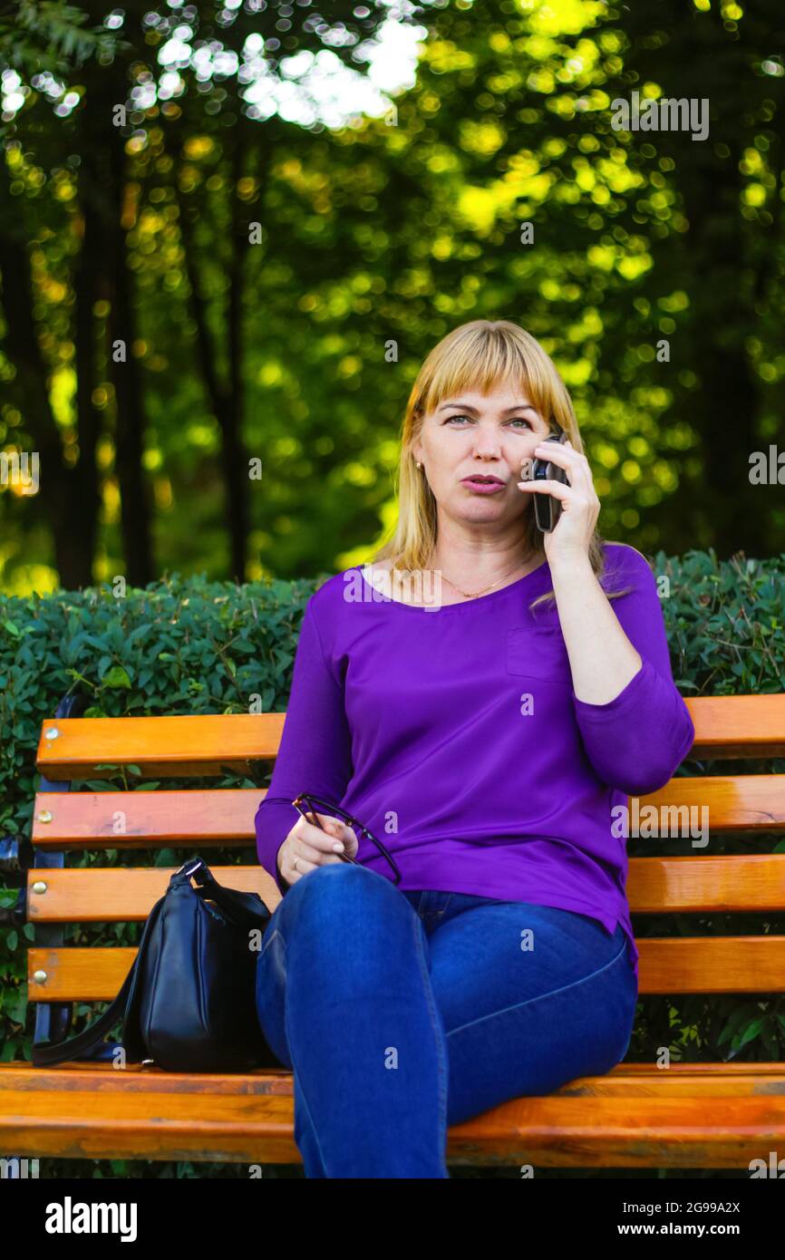 Unschärfe ernsthafte kaukasische blonde Frau sprechen, sprechen am Telefon draußen, im Freien. 40-jährige Frau in lila Bluse im Park. Erwachsene Person Stockfoto