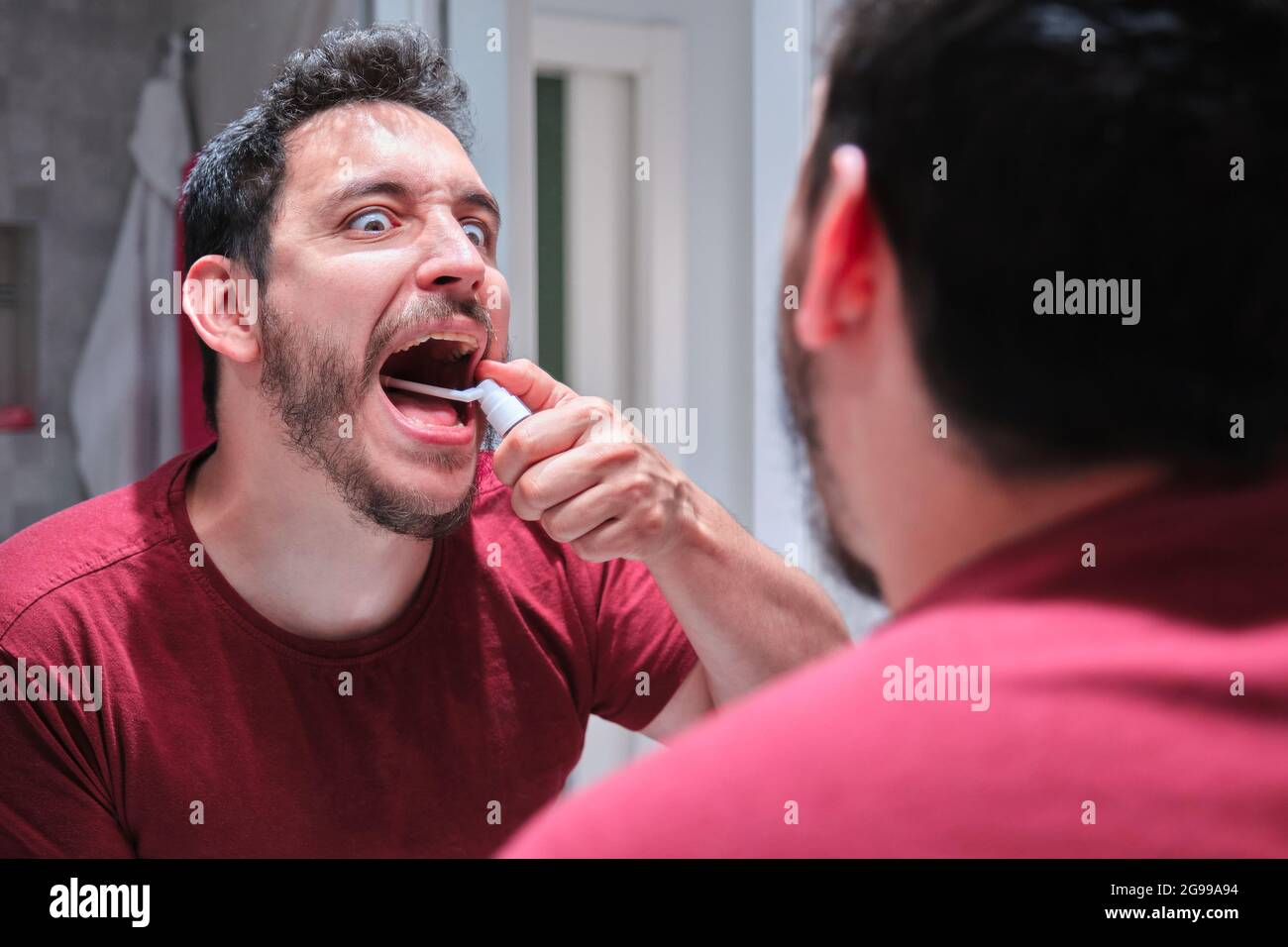 Mann mit Schmerzen, während er ein Spray zur Behandlung von Aphthen oder Halsschmerzen vor dem Spiegel verwendete. Stockfoto
