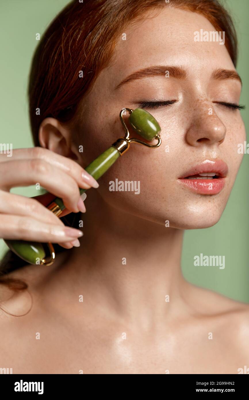 Feminine schöne Frau mit roten Haaren, geschlossenen Augen, massieren  Gesicht mit guasha Jade Roller, Kosmetik-Verfahren zu Hause,  Anti-Aging-Konzept, grün Stockfotografie - Alamy