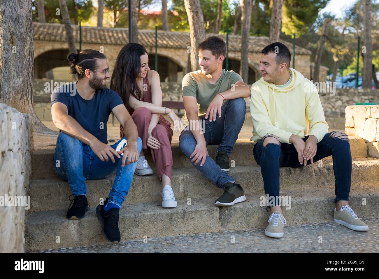 Eine Gruppe von Freunden, die auf der Parktreppe sitzen und lachen. Sie sind kaukasisch und tragen Sommerkleidung. Sie befinden sich in einem Park mit Kiefern. Javea, Alicant Stockfoto